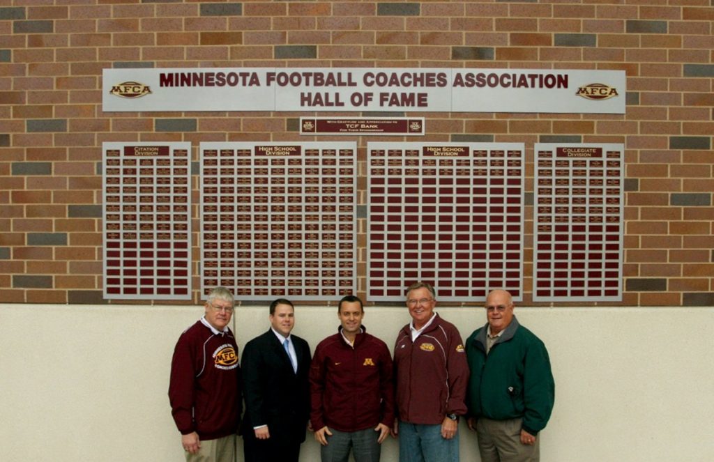 Coaches and staff pose in front of Minnesota Football Coaches Association Hall of Fame